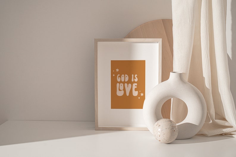 May include: A framed print with the words "GOD IS LOVE" in white on an orange background. A white, circular vase and a decorative sphere sit beside the frame. A light-colored curtain hangs in the background.