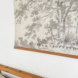 Puede incluir: Una impresi&oacute;n en blanco y negro de un boceto de &aacute;rbol con las palabras "ROOTED & GROUNDED" en la parte inferior. La impresi&oacute;n est&aacute; sujeta por un marco de madera y cuelga de una pared blanca. Un marco de cama de madera y almohadas blancas est&aacute;n en primer plano.