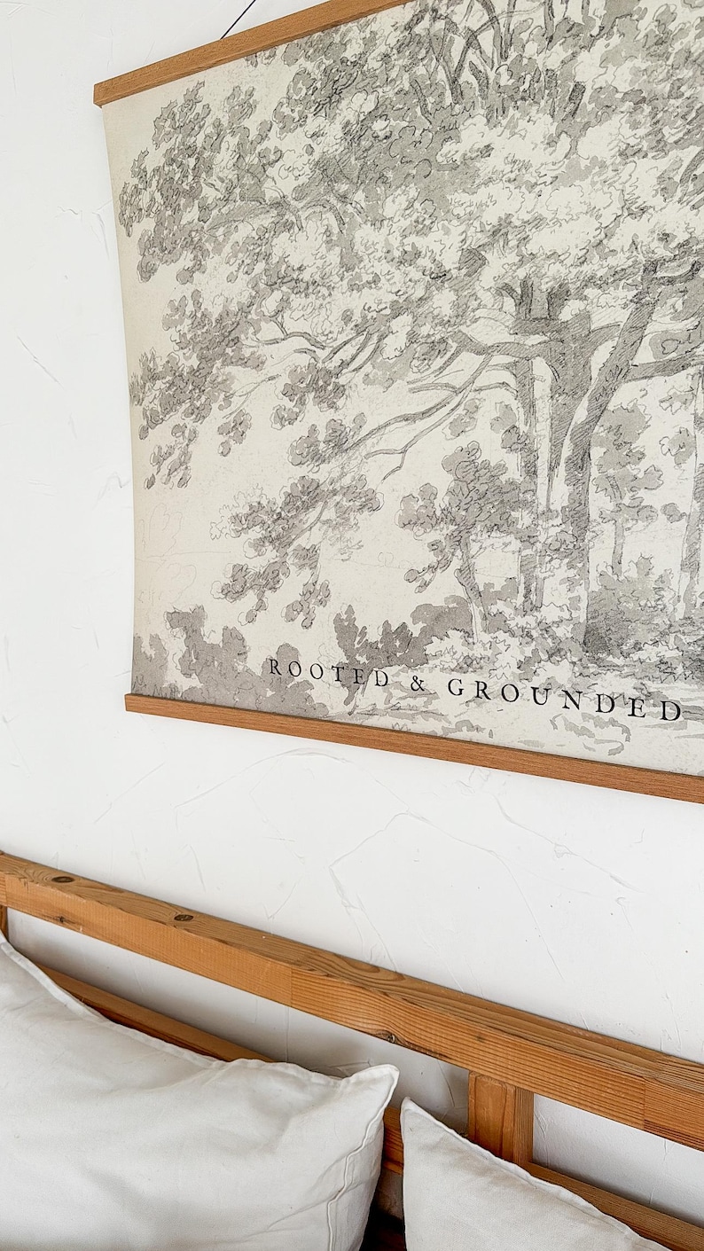 Puede incluir: Una impresi&oacute;n en blanco y negro de un boceto de &aacute;rbol con las palabras "ROOTED & GROUNDED" en la parte inferior. La impresi&oacute;n est&aacute; sujeta por un marco de madera y cuelga de una pared blanca. Un marco de cama de madera y almohadas blancas est&aacute;n en primer plano.