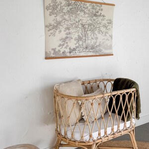 Puede incluir: Una cuna de beb&eacute; de rat&aacute;n marr&oacute;n claro con un dise&ntilde;o tejido, que contiene almohadas y una manta verde. Un tapiz de pared con una impresi&oacute;n de &aacute;rbol y el texto "ROOTED & GROUNDED IN LOVE" est&aacute; encima de la cuna. Una peque&ntilde;a mecedora de madera est&aacute; cerca.