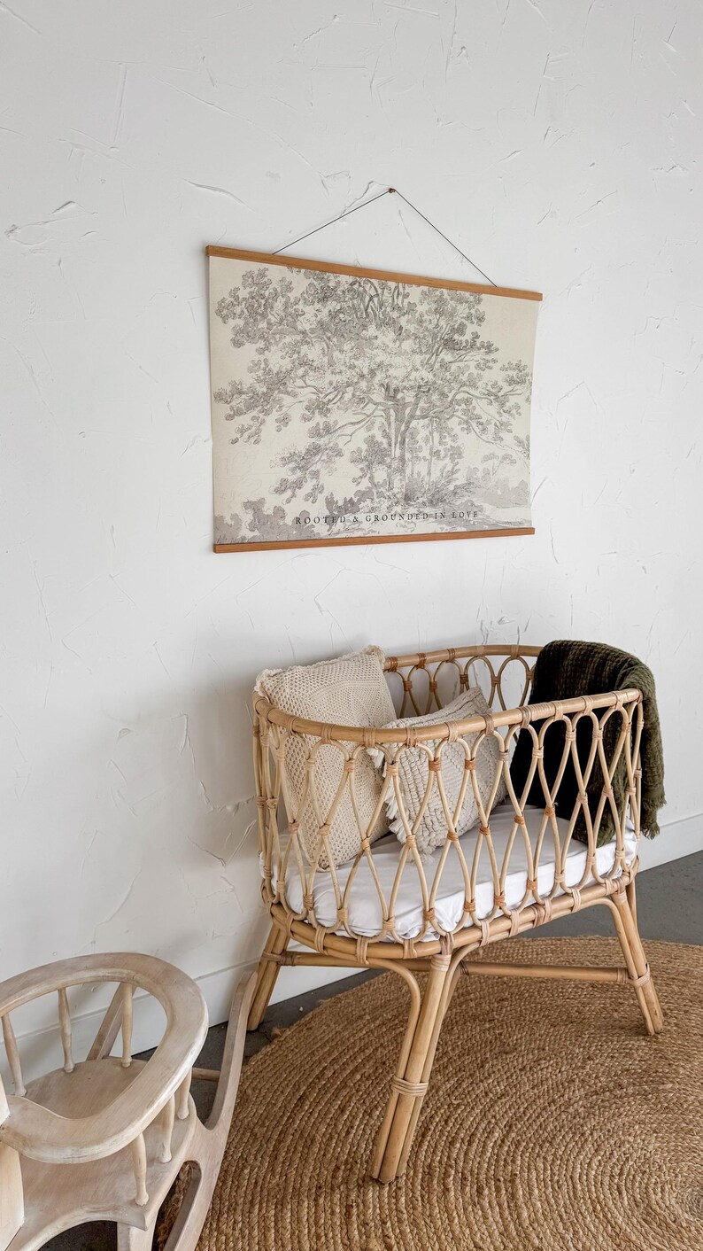 Puede incluir: Una cuna de beb&eacute; de rat&aacute;n marr&oacute;n claro con un dise&ntilde;o tejido, que contiene almohadas y una manta verde. Un tapiz de pared con una impresi&oacute;n de &aacute;rbol y el texto "ROOTED & GROUNDED IN LOVE" est&aacute; encima de la cuna. Una peque&ntilde;a mecedora de madera est&aacute; cerca.