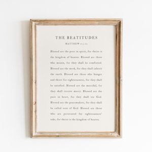 May include: A framed print with the text "THE BEATITUDES" and a passage from Matthew. The text is in a simple, elegant font on an off-white background, framed by a light brown wooden frame. The print is displayed against a white wall.
