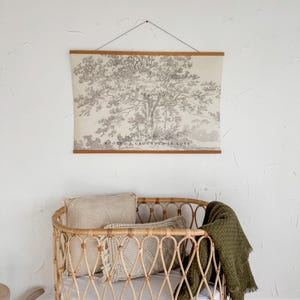 Puede incluir: Una escena de guarder&iacute;a con una cuna de rat&aacute;n marr&oacute;n claro, un colch&oacute;n blanco, almohadas beige y una manta verde oliva. Encima de la cuna, un estampado enmarcado con una ilustraci&oacute;n de &aacute;rbol y el texto "ROOTED & GROUNDED IN LOVE."