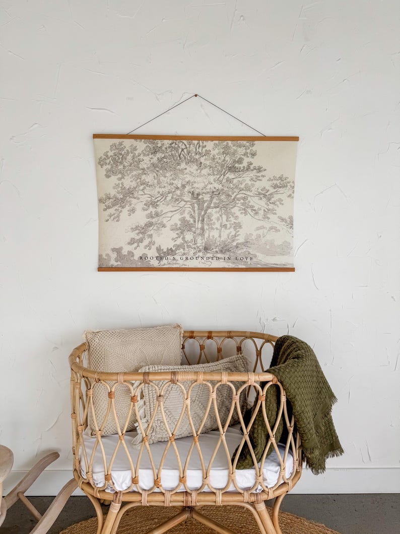 Puede incluir: Una escena de guarder&iacute;a con una cuna de rat&aacute;n marr&oacute;n claro, un colch&oacute;n blanco, almohadas beige y una manta verde oliva. Encima de la cuna, un estampado enmarcado con una ilustraci&oacute;n de &aacute;rbol y el texto "ROOTED & GROUNDED IN LOVE."