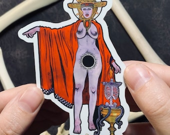 May include: A die-cut sticker featuring a pale figure with a golden headdress and snakes, wearing an orange cloak. A black sun symbol is centered on the torso. Beside the figure is a golden chalice with a skeletal rib cage and purple tentacles.