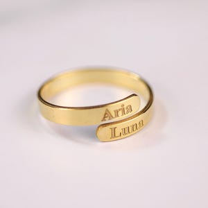 May include: A gold-colored wrap ring with the names "Aria" and "Luna" engraved on the band. The ring is displayed on a white surface, showcasing its minimalist design and personalized detail.