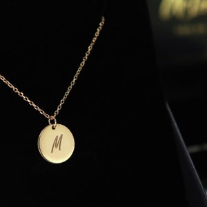Könnte beinhalten: Eine goldene Kettenkette mit einem runden Anhänger, auf dem der Buchstabe "M" eingraviert ist.