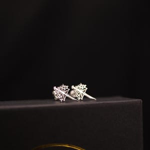 Puede incluir: Un par de pendientes de botón plateados con un diseño de copo de nieve calado. Los pendientes se muestran sobre una superficie oscura sobre un fondo negro. Los pendientes son pequeños y delicados.
