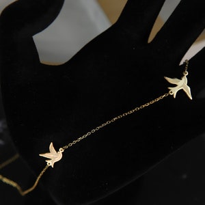 May include: A gold-coloured necklace with two bird-shaped pendants linked by a fine chain. The birds face opposite ways. The necklace is shown against a black backdrop, accentuating its elegant design.