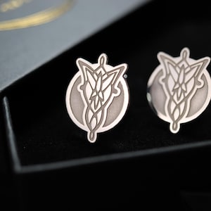 Puede incluir: Un par de gemelos de plata con un diseño grabado en blanco de un árbol estilizado. Los gemelos se muestran en una caja negra.