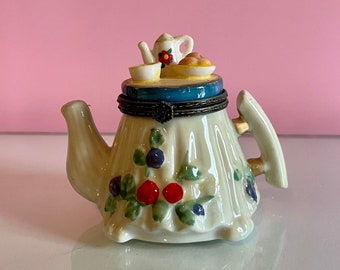 May include: A white porcelain teapot trinket box with a floral design and a small teapot, cup, and plate on top. The teapot has a red flower on the side.