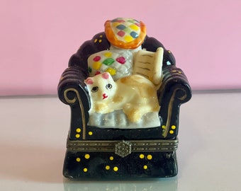 May include: A black and white porcelain figurine of a cat sitting in a black armchair with a colorful patterned pillow. The armchair has gold accents and black polka dots.