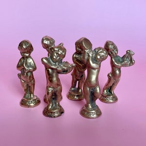 May include: A set of seven small gold-toned figurines of cherubs playing musical instruments. The cherubs are all standing on small bases and are arranged in a semi-circle.