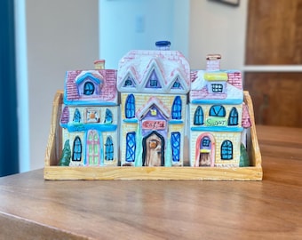 Może przedstawiać: Zestaw czterech ceramicznych pojemników na przyprawy w kształcie domków. Domki są pomalowane w pastelowych kolorach z niebieskimi, żółtymi, różowymi i zielonymi akcentami. Pojemniki są oznaczone jako "Herbata", "Kawa", "Cukier" i "Przyprawy".