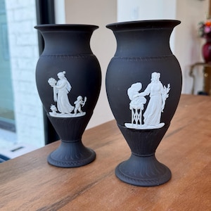 May include: Two matte black ceramic vases featuring white relief figures. The left vase depicts a person holding a basket with a small figure nearby. The right vase shows a person standing by a tripod stand. Both vases have a flared rim and base.