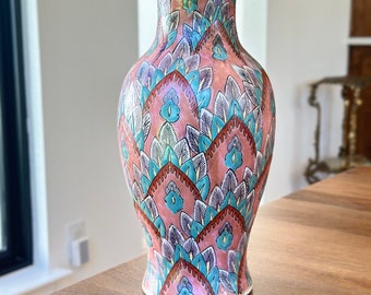 Puede incluir: Un jarrón de cerámica rosa con un patrón floral azul, verde y blanco. El jarrón tiene un cuerpo redondeado y un cuello estrecho.