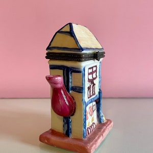 May include: A white ceramic teapot shaped like a house with a pink spout and blue trim. The teapot has a hinged lid and a floral design on the side.