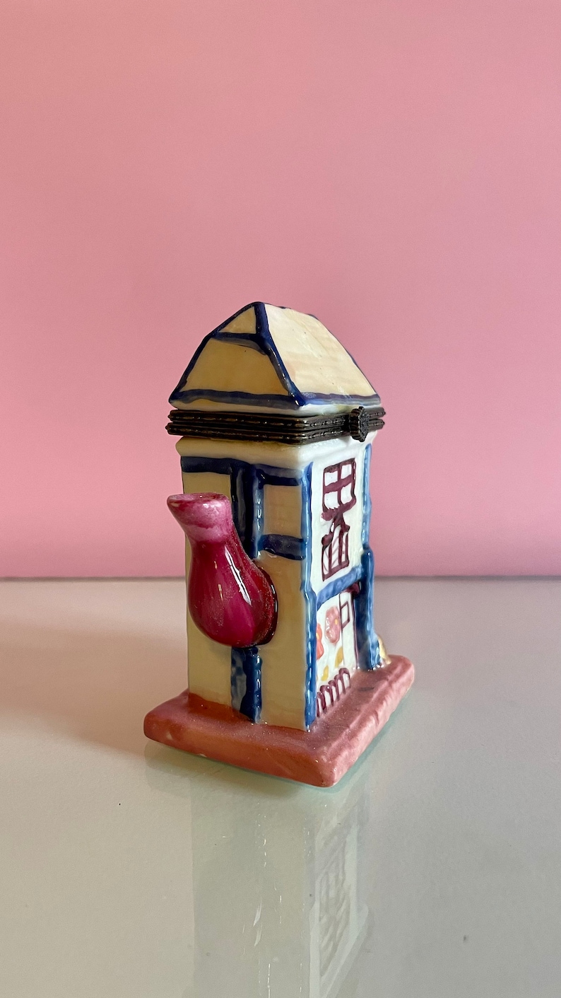May include: A white ceramic teapot shaped like a house with a pink spout and blue trim. The teapot has a hinged lid and a floral design on the side.