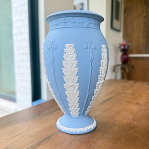 May include: A light blue ceramic vase with white relief detailing. The vase features a flared rim and base, with a series of white leaf and floral patterns adorning the body. The vase is set on a wooden table.