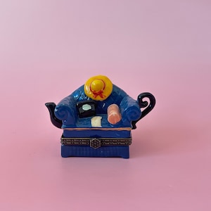 May include: A blue ceramic teapot shaped like a couch with a yellow hat, a book, and a rolled-up blanket on top. The teapot has a black handle and spout.