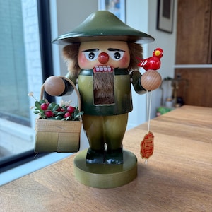 May include: A wooden nutcracker figurine in shades of green, brown, and tan. The figurine holds a basket of berries and a red bird. It has a green hat and a fur-like chest. A tag hangs from the bird's wooden perch.