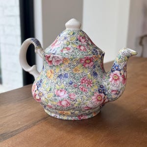 May include: A floral-patterned teapot with a white handle, spout, and lid. The teapot features a colorful design of pink, yellow, blue, and purple flowers on a cream background. The teapot is sitting on a wooden surface.
