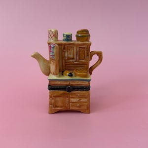Peut inclure: Une théière en céramique en forme de miniature de cuisine avec un schéma de couleurs brun, crème et rouge. La théière a un bec verseur et une poignée et présente une petite fenêtre avec un motif à carreaux rouge et blanc. L'armoire a une petite étagère avec un motif brun et blanc.