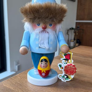 May include: A wooden nutcracker in the shape of a man with a blue coat, hat, and pants, and a white beard. A small Matryoshka doll and a tag are next to the nutcracker. The nutcracker is on a wooden surface.