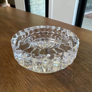 May include: A clear, circular glass ashtray featuring a heavy, faceted design with vertical cut patterns along the exterior rim and a starburst pattern etched into the center base, creating a reflective and textured appearance throughout the piece.