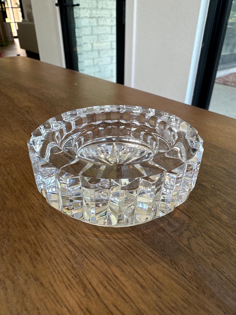 May include: A clear, circular glass ashtray featuring a heavy, faceted design with vertical cut patterns along the exterior rim and a starburst pattern etched into the center base, creating a reflective and textured appearance throughout the piece.