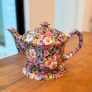 Könnte beinhalten: Eine dekorative Teekanne mit floralem Muster. Die Teekanne hat einen schwarzen Hintergrund, verziert mit einer lebendigen Mischung aus rosa, gelben und weißen Blumen. Deckel, Henkel und Ausguss sind ebenfalls mit dem gleichen floralen Design verziert.