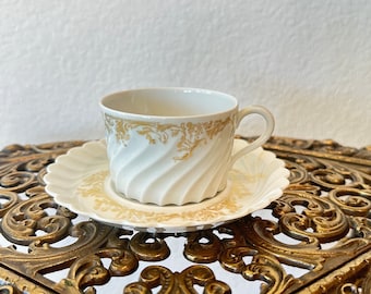 May include: A white teacup and saucer with a gold floral pattern. The cup and saucer have a swirl design and are sitting on a gold metal stand.