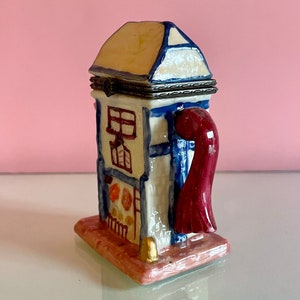 May include: A ceramic trinket box shaped like a small house with a red chimney and a blue door. The house is painted with white and yellow accents and has a small window. The box is sitting on a pink base.