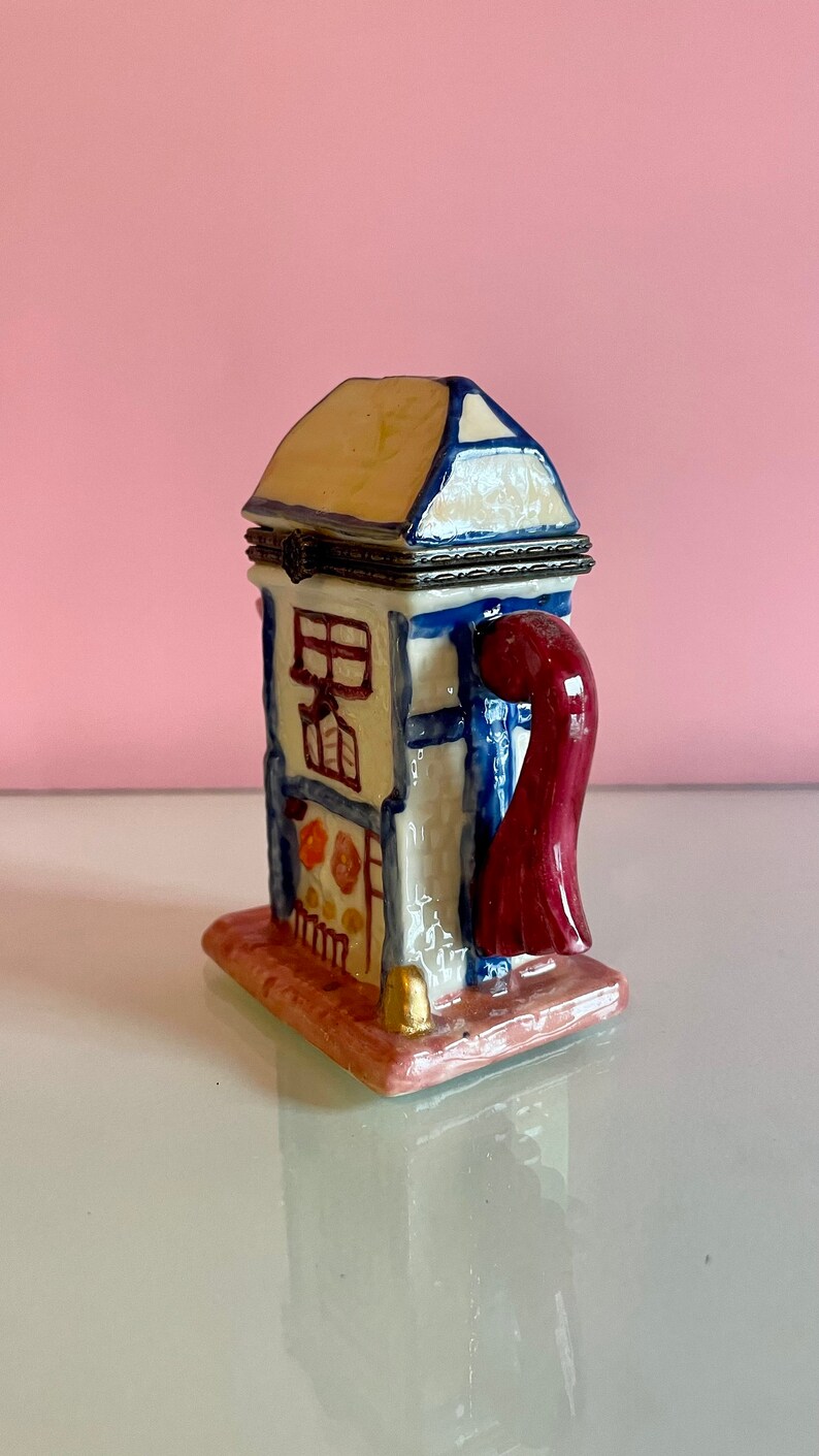 May include: A ceramic trinket box shaped like a small house with a red chimney and a blue door. The house is painted with white and yellow accents and has a small window. The box is sitting on a pink base.