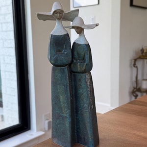 May include: Two ceramic figurines of nuns standing side-by-side. They are wearing blue and green robes and white head coverings. The figurines are on a wooden surface.