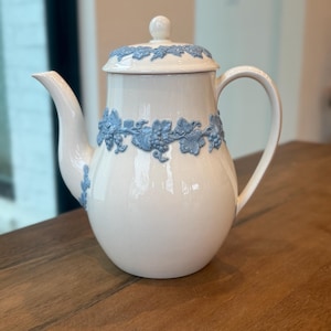 May include: A white ceramic teapot with a rounded body, a curved spout, and a large handle. The teapot features a blue grape vine design around the body and on the lid. The lid has a small, round knob. The teapot is sitting on a wooden surface.