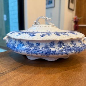 May include: A white and blue ceramic serving dish with a lid. The dish features a floral pattern in blue and gold trim. The dish has a handle on the lid and two handles on the sides.