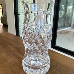 May include: A clear, colorless glass vase featuring a flared rim and a body decorated with a deep, diamond-cut geometric pattern. The base is thick and circular, also adorned with a series of vertical, notched cuts around its outer edge.
