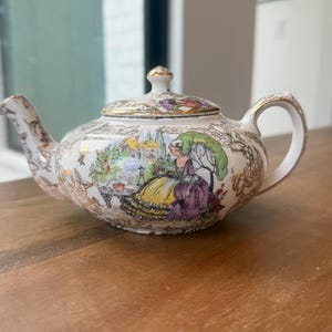 Puede incluir: Una tetera de porcelana blanca con una ilustración colorida de una persona con un vestido amarillo y morado sentada en un jardín. La tetera tiene una tapa con borde dorado, un asa curva, un pico y motivos florales dorados en el cuerpo.