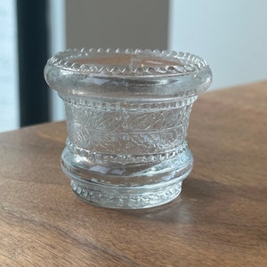Può includere: Un portacandele in vetro trasparente con un motivo decorativo a foglie. Il portacandele ha una base rotonda e una parte superiore svasata con un bordo perlato. Il vetro è trasparente, permettendo alla luce di passare.