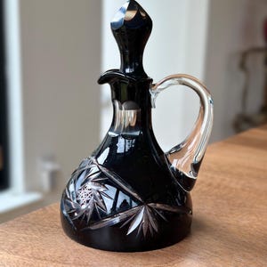 May include: A black glass cruet with a clear glass handle. The cruet features a faceted stopper and a cut glass design on the base. The cruet is sitting on a wooden surface.