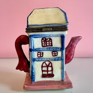 May include: A white ceramic teapot shaped like a house with a blue roof and red trim. The teapot has a red handle and spout. The house has windows and a door.