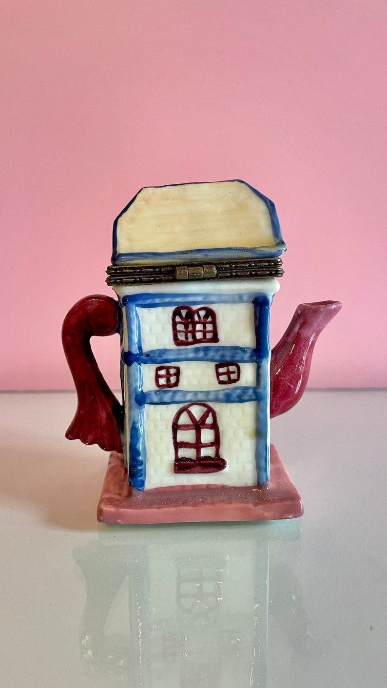 May include: A white ceramic teapot shaped like a house with a blue roof and red trim. The teapot has a red handle and spout. The house has windows and a door.