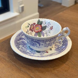 May include: A vintage teacup and saucer set. The teacup features a floral design with red and purple flowers. The saucer and cup have a blue and white pattern. The rim of the saucer has a scalloped edge.