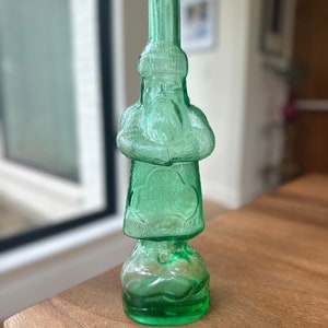 Pode incluir: Uma garrafa de vidro verde em forma de figura de Papai Noel. A garrafa tem uma superfície texturizada e uma base redonda.