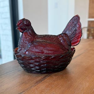Pode incluir: Um prato de vidro vermelho rubi em forma de galinha com uma superfície texturizada. A galinha está sentada numa base em forma de cesta tecida. As penas da cauda estão abertas. Esta peça decorativa é adequada para decoração de cozinha.