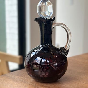 May include: A dark purple glass decanter with a clear glass stopper and etched grape design. The decanter has a round body and a short neck with a handle.