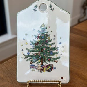 May include: White ceramic Christmas tree cutting board with a green border. The board features a green Christmas tree with ornaments, presents, and a teddy bear. The text "Noritake Company Japan" is printed on the bottom right corner.