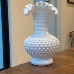 May include: A white milk glass vase with a ruffled top and a textured, dotted surface. The vase is sitting on a wooden table.