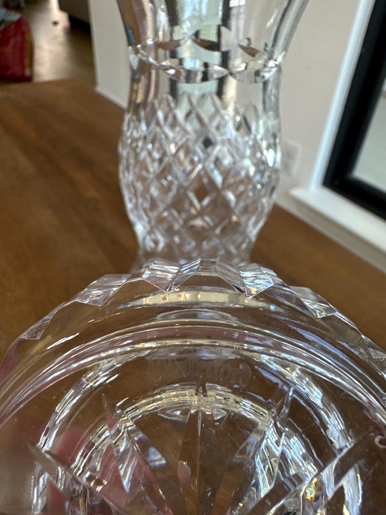May include: A clear glass vase featuring a prominent diamond-cut pattern across its body and a flared rim. The base of the vase is circular with a starburst design etched into the bottom, showing the Waterford brand name clearly visible in the glass.